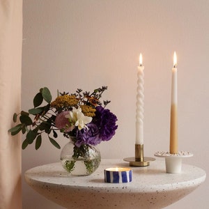 May include: A white table with a speckled surface, featuring a bouquet of flowers in a clear glass vase, two lit candles in gold holders, and a blue and white striped candle.