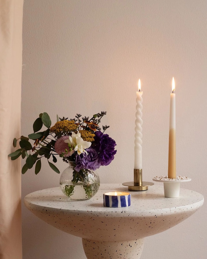 May include: A white table with a speckled surface, featuring a bouquet of flowers in a clear glass vase, two lit candles in gold holders, and a blue and white striped candle.
