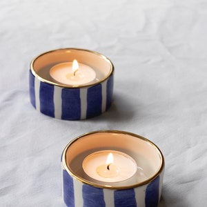 May include: Two white ceramic candle holders with blue and gold stripes. Each holder has a lit tea light candle inside.