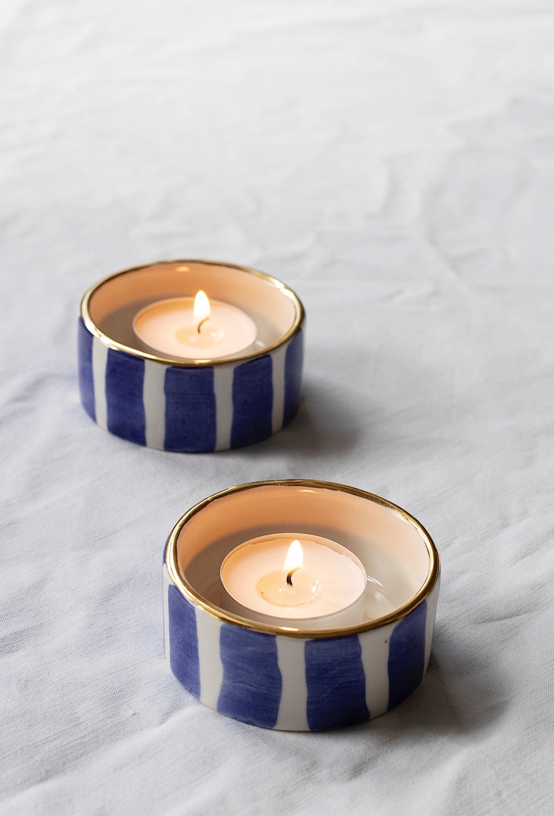 May include: Two white ceramic candle holders with blue and gold stripes. Each holder has a lit tea light candle inside.
