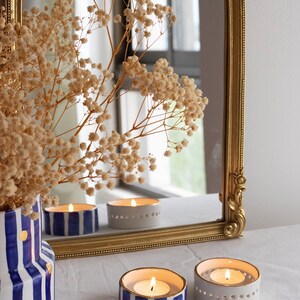May include: Three white ceramic candle holders with gold trim. Two holders have blue and gold stripes and one is plain white. The candle holders are lit and the flames are visible. The candle holders are on a white surface in front of a gold framed mirror.