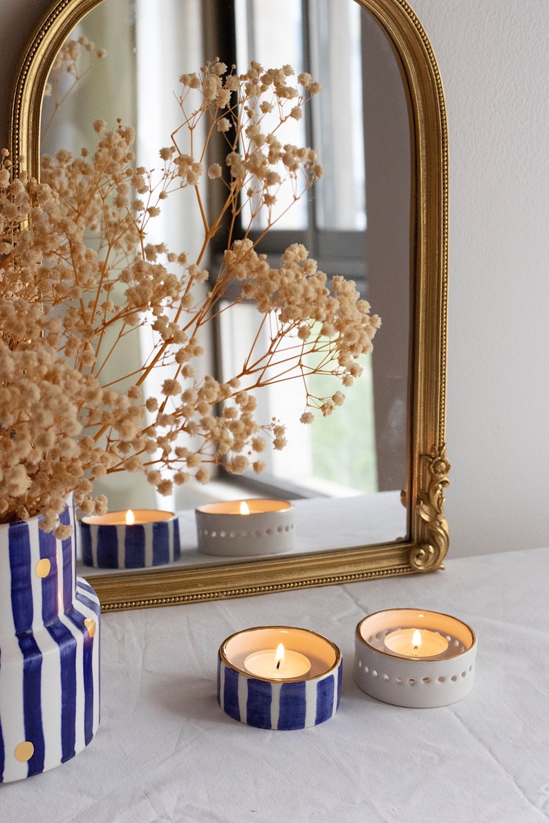 May include: Three white ceramic candle holders with gold trim. Two holders have blue and gold stripes and one is plain white. The candle holders are lit and the flames are visible. The candle holders are on a white surface in front of a gold framed mirror.
