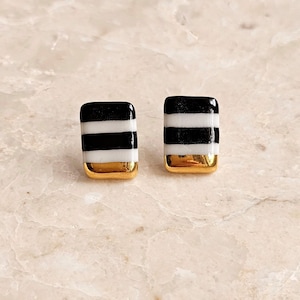 Op de afbeelding: Een paar vierkante oorstekers met een zwart-wit gestreept ontwerp. De onderkant van elke oorbel is goudkleurig. De oorbellen worden gepresenteerd op een lichtgekleurd, marmerachtig oppervlak.