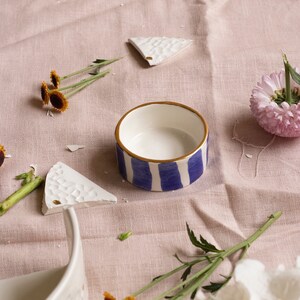 May include: A white ceramic bowl with blue and gold stripes sits on a pink linen tablecloth. The bowl is surrounded by small white ceramic pieces, yellow flowers, and a white flower.