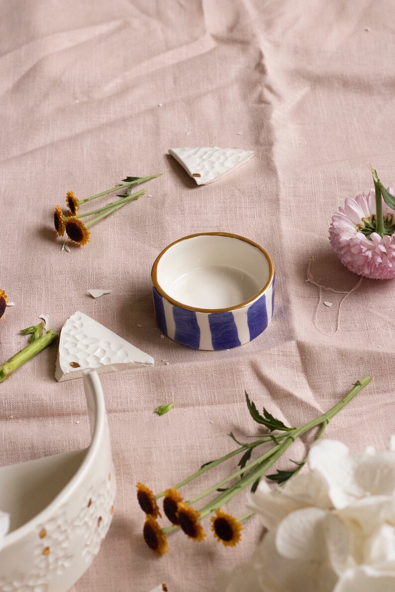 May include: A white ceramic bowl with blue and gold stripes sits on a pink linen tablecloth. The bowl is surrounded by small white ceramic pieces, yellow flowers, and a white flower.