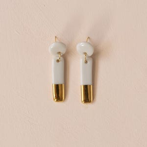Puede incluir: Un par de pendientes colgantes que presentan un pequeño poste circular blanco conectado a un colgante rectangular blanco. La mitad inferior de cada colgante rectangular tiene un acabado con un revestimiento de oro metálico y las piezas están unidas por anillas de metal dorado.