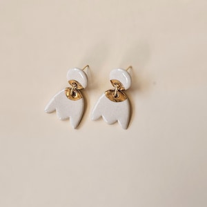 Peut inclure: Une paire de boucles d'oreilles en céramique blanche avec des accents dorés. Chaque boucle d'oreille a un dessus arrondi et un bas stylisé à trois lobes. Les boucles d'oreilles sont présentées sur un fond neutre, soulignant leur design minimaliste.
