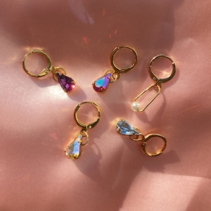 Könnte beinhalten: Fünf goldene Creolen mit baumelnden tropfenförmigen Kristallen. Die Kristalle sind irisierend und haben einen Regenbogeneffekt. Die Ohrringe liegen auf einem rosa Hintergrund.