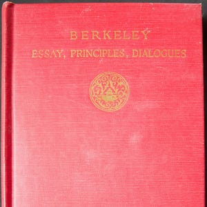 May include: A red book with gold lettering that reads "BERKELEY ESSAY, PRINCIPLES, DIALOGUES". A gold circular emblem is centered below the text. The book has a textured cover.