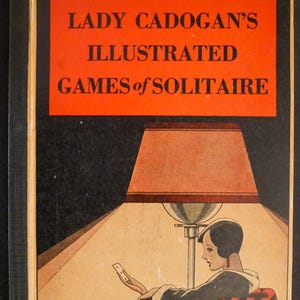 May include: Vintage book cover titled "Lady Cadogan's Illustrated Games of Solitaire." The cover features an illustration of a woman seated in a patterned chair, playing cards under a lamp. The background is black with an orange banner.