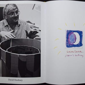 May include: Black and white photograph of David Hockney with a striped shirt, looking at a cylindrical cardboard structure. On the facing page, a drawing of a moon in a square with the text "Luca Luna" and "Sam Whitney".