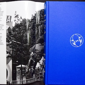 May include: A hardcover book with a bright blue cover and a white circular emblem. The book is open, revealing black and white images and text. The text on the left page describes the history of Luna Luna, the first art amusement park.