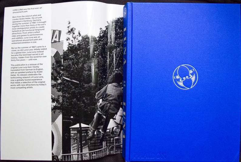 May include: A hardcover book with a bright blue cover and a white circular emblem. The book is open, revealing black and white images and text. The text on the left page describes the history of Luna Luna, the first art amusement park.