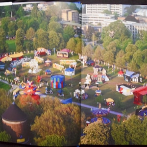May include: Aerial view of a fairground with colorful rides and booths set amidst lush green trees. Buildings and a cityscape are visible in the background, creating a vibrant scene.