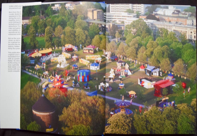 May include: Aerial view of a fairground with colorful rides and booths set amidst lush green trees. Buildings and a cityscape are visible in the background, creating a vibrant scene.
