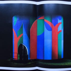 May include: An abstract architectural illustration featuring a cylindrical structure with a dark arched doorway. The structure is painted with vertical stripes of blue, green, and red, with red tree-like shapes. The background is black, and the image is on a book page.