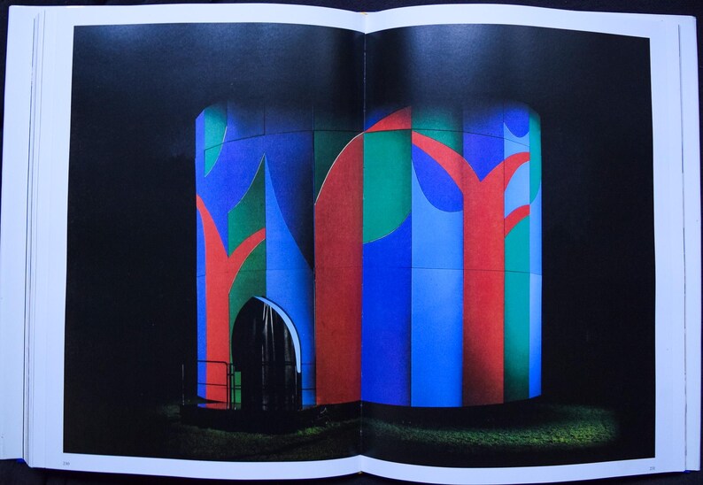 May include: An abstract architectural illustration featuring a cylindrical structure with a dark arched doorway. The structure is painted with vertical stripes of blue, green, and red, with red tree-like shapes. The background is black, and the image is on a book page.