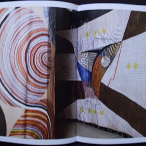May include: An open book displaying abstract art. One page features a spiral design in shades of brown, orange, and white. The other page shows geometric shapes with white, brown, and blue elements, accented with yellow dots.