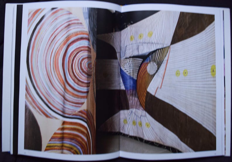 May include: An open book displaying abstract art. One page features a spiral design in shades of brown, orange, and white. The other page shows geometric shapes with white, brown, and blue elements, accented with yellow dots.