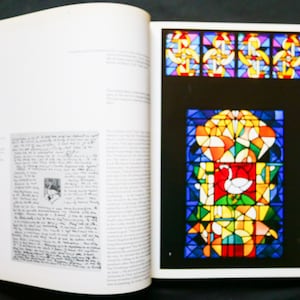 May include: Open book with a red spine, displaying a page with handwritten text and a page with colorful stained glass art. The stained glass features geometric shapes and a swan design in red, white, and green.