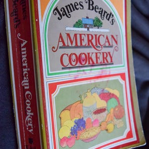 May include: A vintage cookbook titled "James Beard's American Cookery" with a colorful illustration of food. The cover features a house illustration and the title in red and black lettering. The book's spine is red.