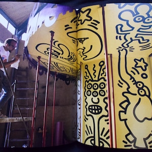 May include: An open book featuring a yellow wall with black line art. A person on a ladder is painting the wall. The art includes a smiling moon, stars, and various cartoon figures. The overall theme is art and design.