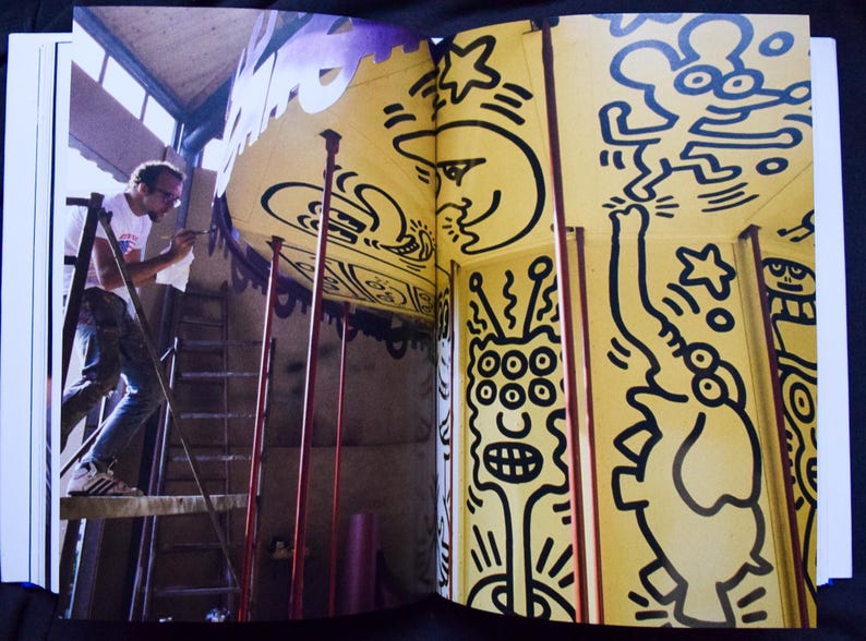 May include: An open book featuring a yellow wall with black line art. A person on a ladder is painting the wall. The art includes a smiling moon, stars, and various cartoon figures. The overall theme is art and design.