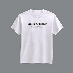 Puede incluir: Camiseta blanca de cuello redondo con el texto "ACHY & TIRED" en negrita oscura, con "Rheumatoid Arthritis" debajo en una fuente más pequeña. La camiseta se muestra sobre un fondo gris.