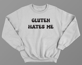 May include: A light gray sweatshirt with the text "GLUTEN HATES ME" in bold, black, rounded letters. The sweatshirt has a crew neck and long sleeves, and is made of a soft material.