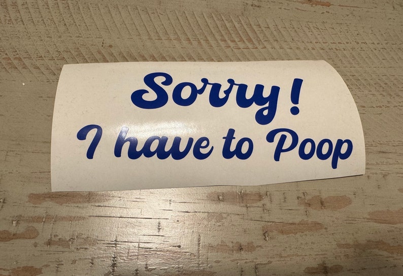 Może przedstawiać: Biała naklejka winylowa z napisem "Sorry! I have to Poop" w niebieskiej, kursywnej czcionce. Naklejka ma zakrzywiony kształt i jest umieszczona na jasnej drewnianej powierzchni.