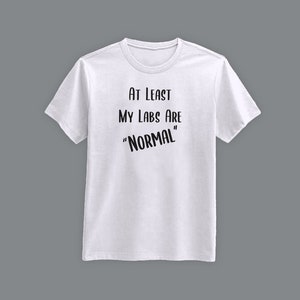 May include: White crew neck t-shirt with the text "AT LEAST MY LABS ARE 'NORMAL'" in black. The shirt is displayed on a grey background. The text is in a casual, handwritten style.