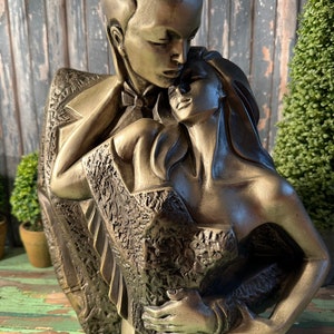 May include: A bronze sculpture of a couple embracing. The man is wearing a suit and the woman is wearing a dress. The sculpture is detailed and realistic.