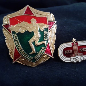 May include: Two vintage enamel pins. The first is a gold-toned shield with a red star, a green background, and the text "МОТОСТРЕЛКОВЫЕ ВОЙСКА СССР". The second is a white and red enamel pin with the text "XXII 1980" and a depiction of the Olympic rings.