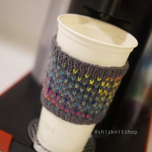 May include: A white paper coffee cup with a gray knitted cozy featuring a colorful pattern of small stitches. The cup is placed on a coffee machine with a red top. The text "@shizknitshop" is visible.