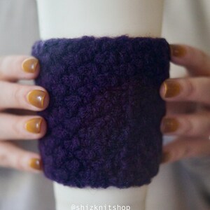 May include: A white disposable coffee cup with a purple knitted cozy. The cozy is a deep purple color and appears to be made of a thick, textured yarn. The cozy is wrapped around the middle of the cup. The shop's Instagram handle, @shizknitshop, is visible.