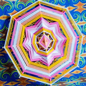 Peut inclure: Un attrape-rêves coloré et multicouche avec un motif rose, blanc et jaune. L'attrape-rêves est fait de fil et a un centre en forme d'étoile.