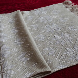 May include: A beige woven table runner with a repeating white snowflake pattern. The runner has a fringed edge and is folded over, revealing the intricate design. The runner is displayed on a red surface.