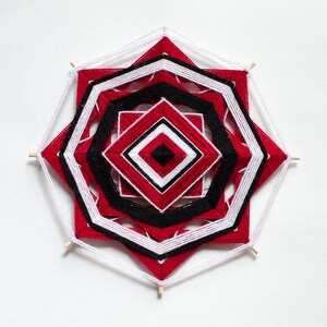 Op de afbeelding: Een rode, witte en zwarte garenmandala met een geometrisch patroon. De mandala is gemaakt van touw en hangt aan een witte muur.
