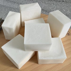 Può includere: Una collezione di saponette bianche di forma quadrata disposte su un tagliere di legno. Le saponette hanno un aspetto liscio e solido. Un rametto di lavanda secca è visibile sul lato sinistro. Lo sfondo è un materiale argentato lucido.