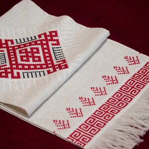 May include: A white linen table runner with red and black embroidered geometric patterns. The runner has a fringe on the bottom edge.