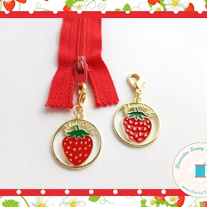 May include: A red zip with two gold charms, each with a red strawberry design and green leaves. The charms have a gold border. The background is white with a red border. The image also includes a second charm with the same design.