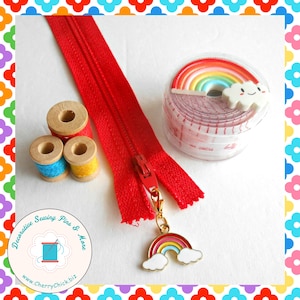 May include: A red zipper, spools of thread, and a rainbow-themed tape measure are displayed. A rainbow charm hangs from the zipper pull. The spools have red, blue, and yellow thread. The tape measure is in a clear container.