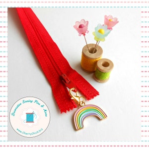 May include: A bright red zipper with a gold-colored rainbow charm, next to two spools of thread and flower-shaped pins. The image also includes a logo with the text "Decorative Sewing Pins & More" and the website address.