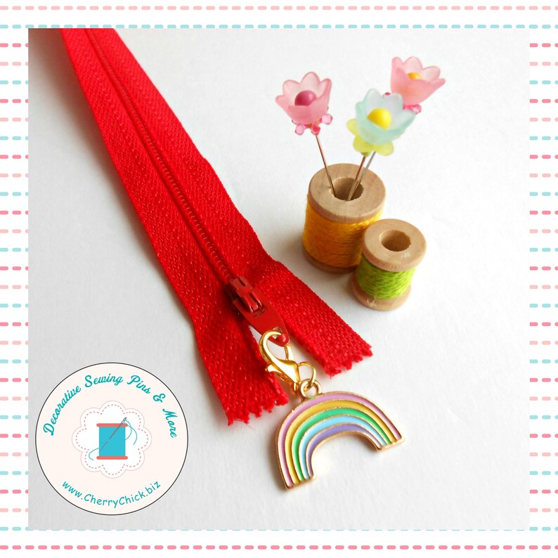 May include: A bright red zipper with a gold-colored rainbow charm, next to two spools of thread and flower-shaped pins. The image also includes a logo with the text "Decorative Sewing Pins & More" and the website address.