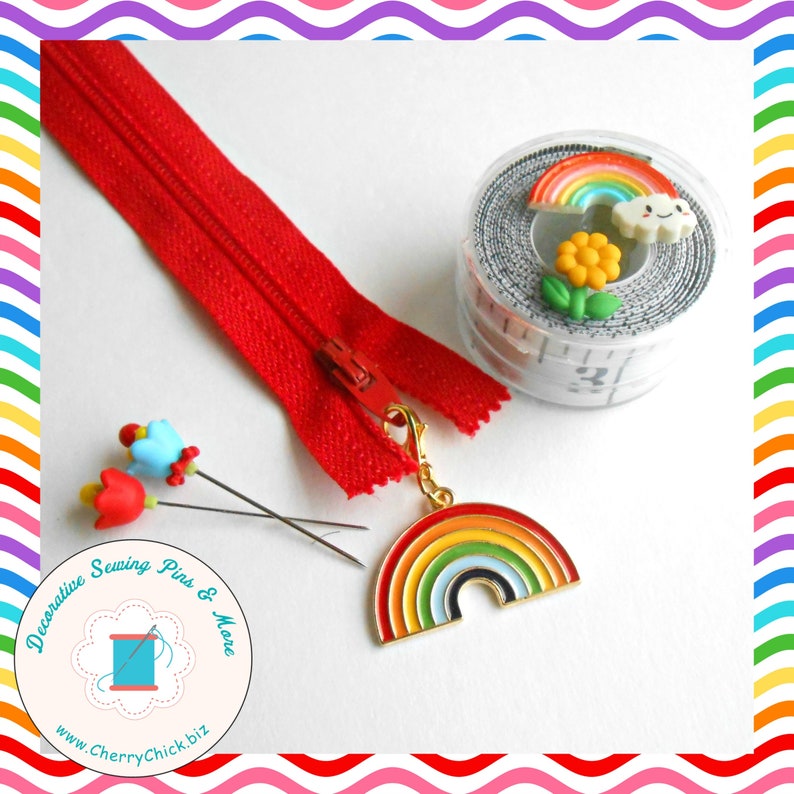 May include: A red zipper, rainbow charm, decorative sewing pins, and a measuring tape with rainbow and flower embellishments are displayed on a white surface. The rainbow charm is multi-colored with a gold-colored clasp.