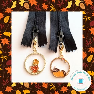 May include: Two black zippers featuring gold-toned metal charms attached with lobster clasps. One charm is a circular frame containing orange and yellow maple leaves, while the other is a circular frame containing two orange and brown acorns.