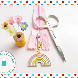 May include: A collection of sewing supplies including a pink zipper, floral-patterned scissors, and a rainbow charm. Decorative flower-shaped pins are displayed in a yellow thread spool. The image also features a logo with the text "Decorative Sewing Pins & More".