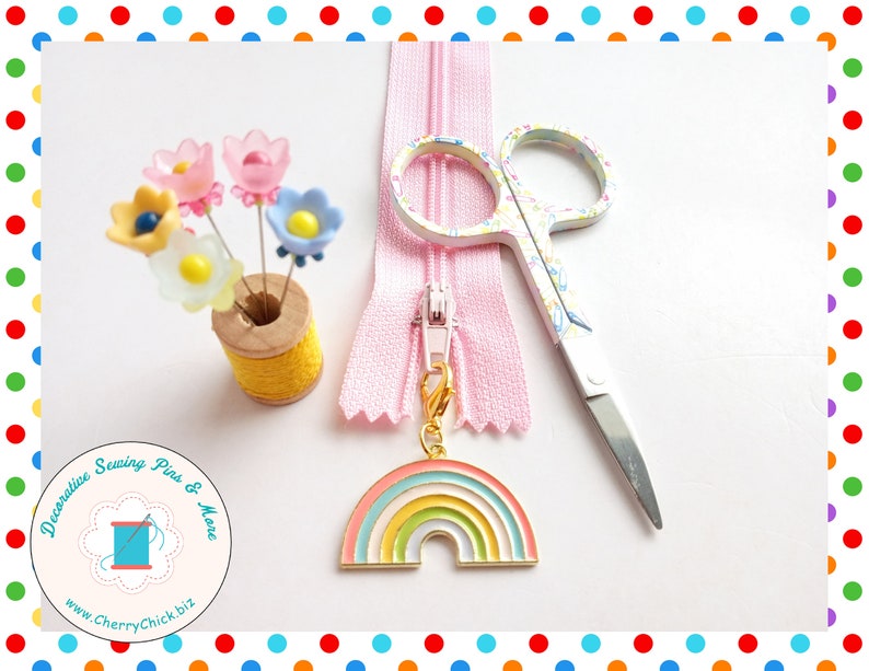 May include: A collection of sewing supplies including a pink zipper, floral-patterned scissors, and a rainbow charm. Decorative flower-shaped pins are displayed in a yellow thread spool. The image also features a logo with the text "Decorative Sewing Pins & More".