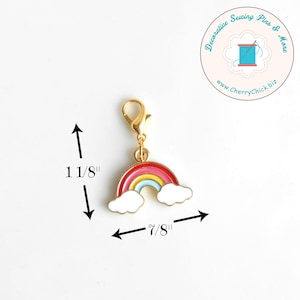 May include: A gold-colored charm with a rainbow design, featuring red, pink, yellow, and blue arches, and white cloud accents. The charm is 1 1/8 inches tall and 7/8 inches wide. It has a lobster clasp for easy attachment. The charm is from Decorative Sewing Pins & More.