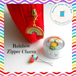 May include: A red zipper pouch with a gold rainbow charm attached. The charm is multi-colored. A measuring tape with a rainbow and flower embellishment is next to the pouch. The text "Rainbow Zipper Charm" is below the pouch.